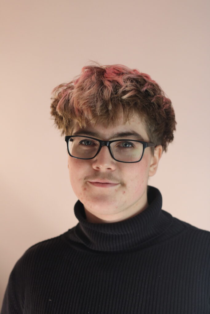 A portrait of Leo in front of a light pink wall, matching their light pink short hair. They are wearing a black elegant turtle neck pullover.