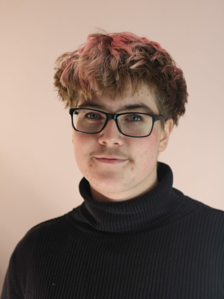 A portrait of Leo in front of a light pink wall, matching their light pink short hair. They are wearing a black elegant turtle neck pullover.