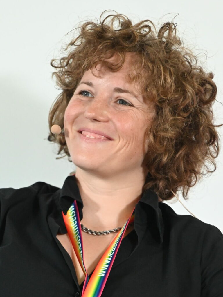 Tabea Hässler smiles in the camera and is wearing a microphone and a badge from the East meets West conference.