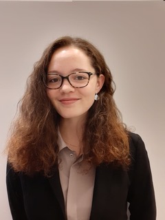 A portrait of Debra wearing black round glasses and long curly red hair in formal clothes smiling at the camera.